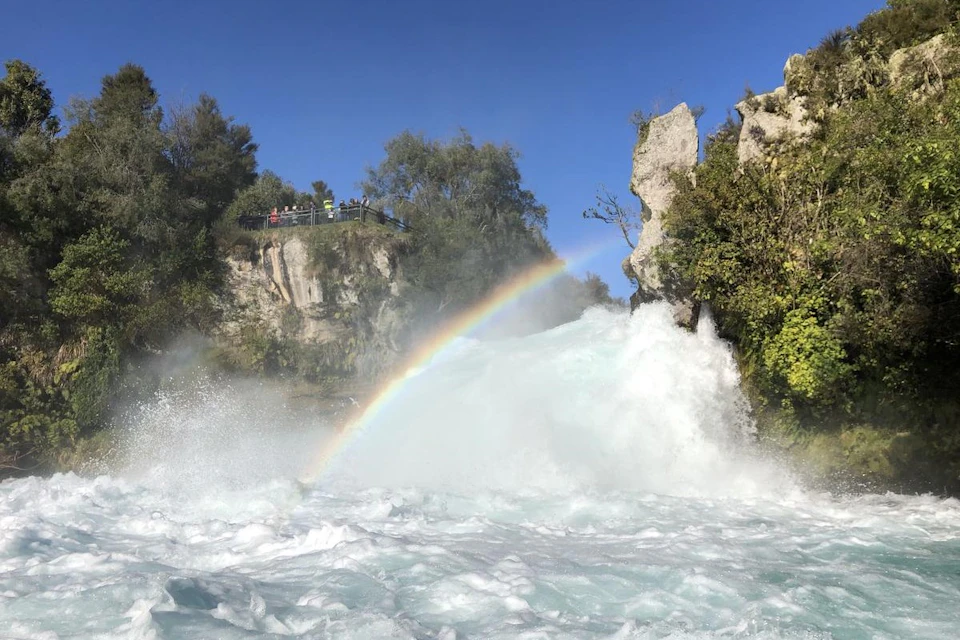 A waterfall with a rainbow in the mist, surrounded by rocky cliffs and greenery under a clear blue sky.