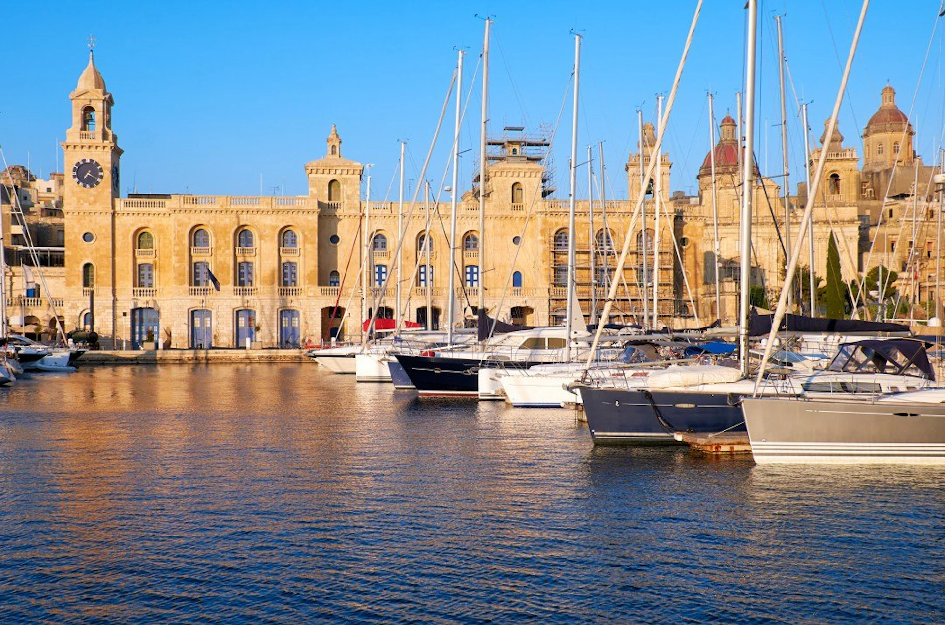 Sailboats docked in a marina with a historic building and church towers in the background under a clear blue sky.