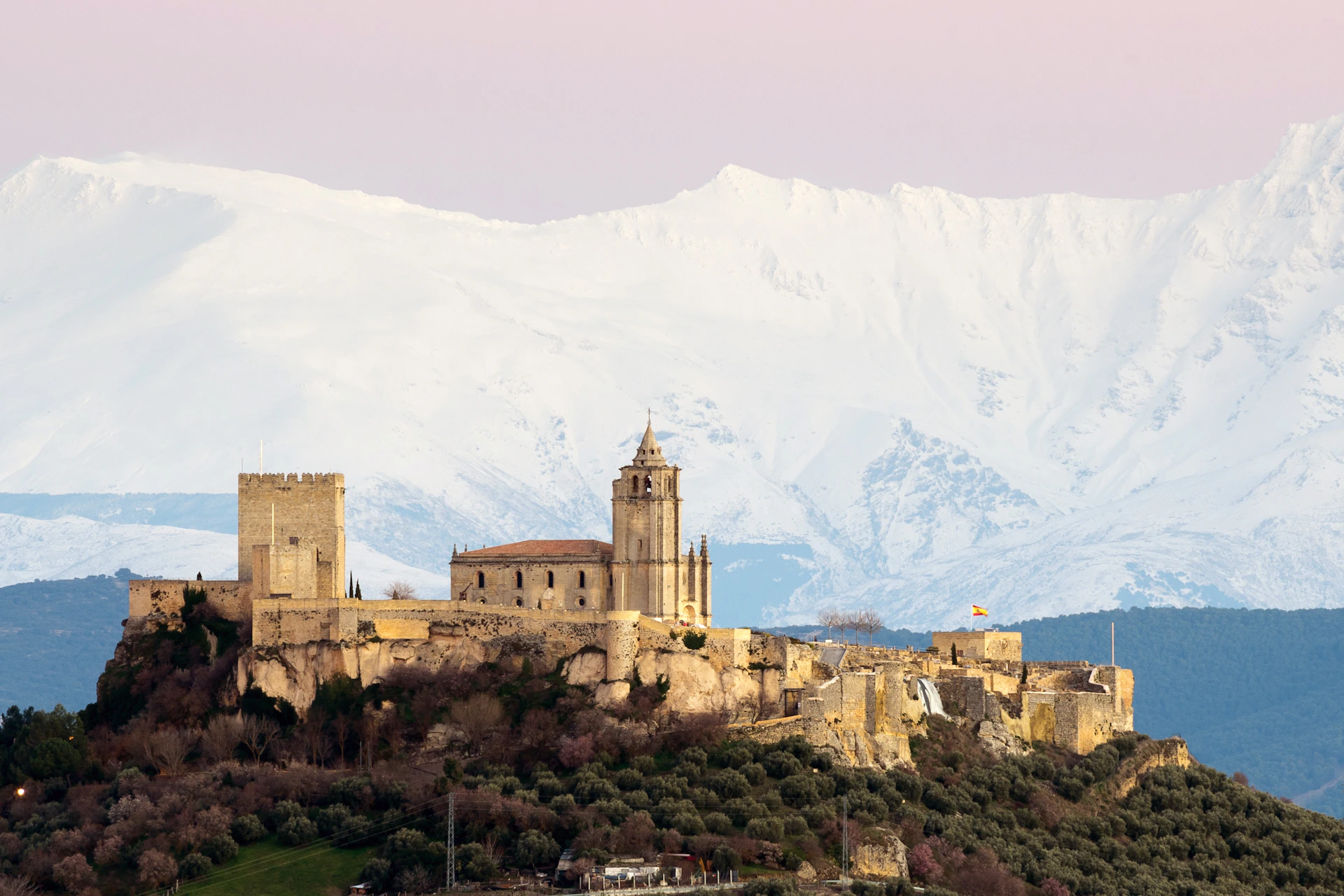 Medieval castle and church on a hilltop with snow-covered mountains in the background under a pinkish sky.