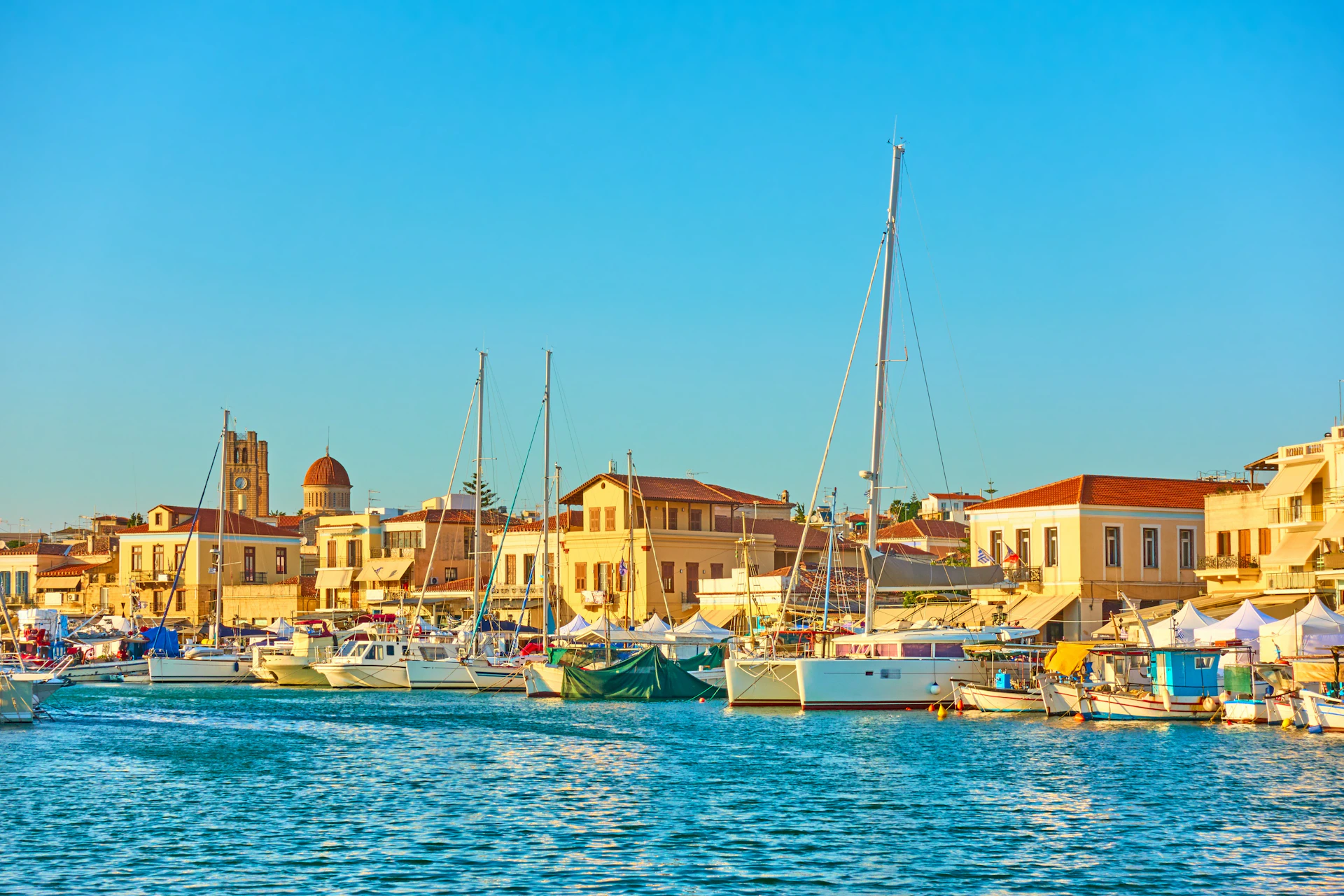 Boats anchored at a harbor with a coastal town and buildings in the background under a clear blue sky.
