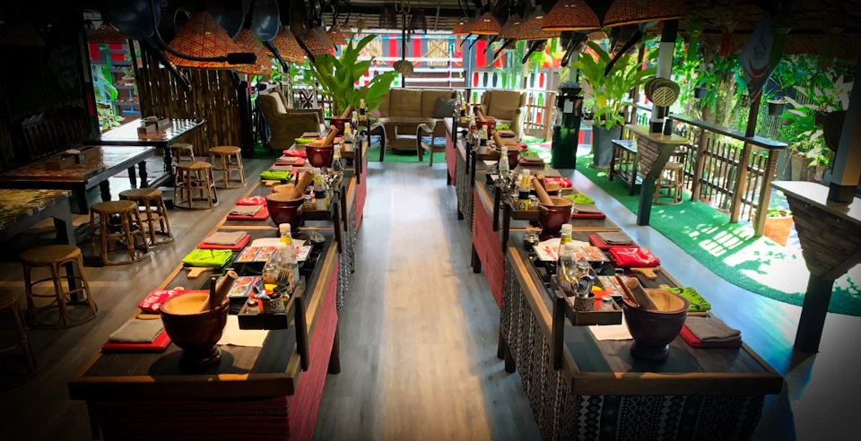 A restaurant with long tables set with utensils, drinks, and condiments, surrounded by plants and decorative lighting.