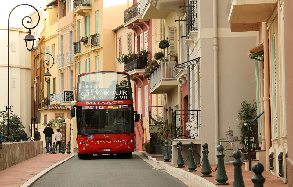 Red double-decker tour bus passing through a narrow street in Monaco, lined with colorful buildings and balconies with potted plants.
