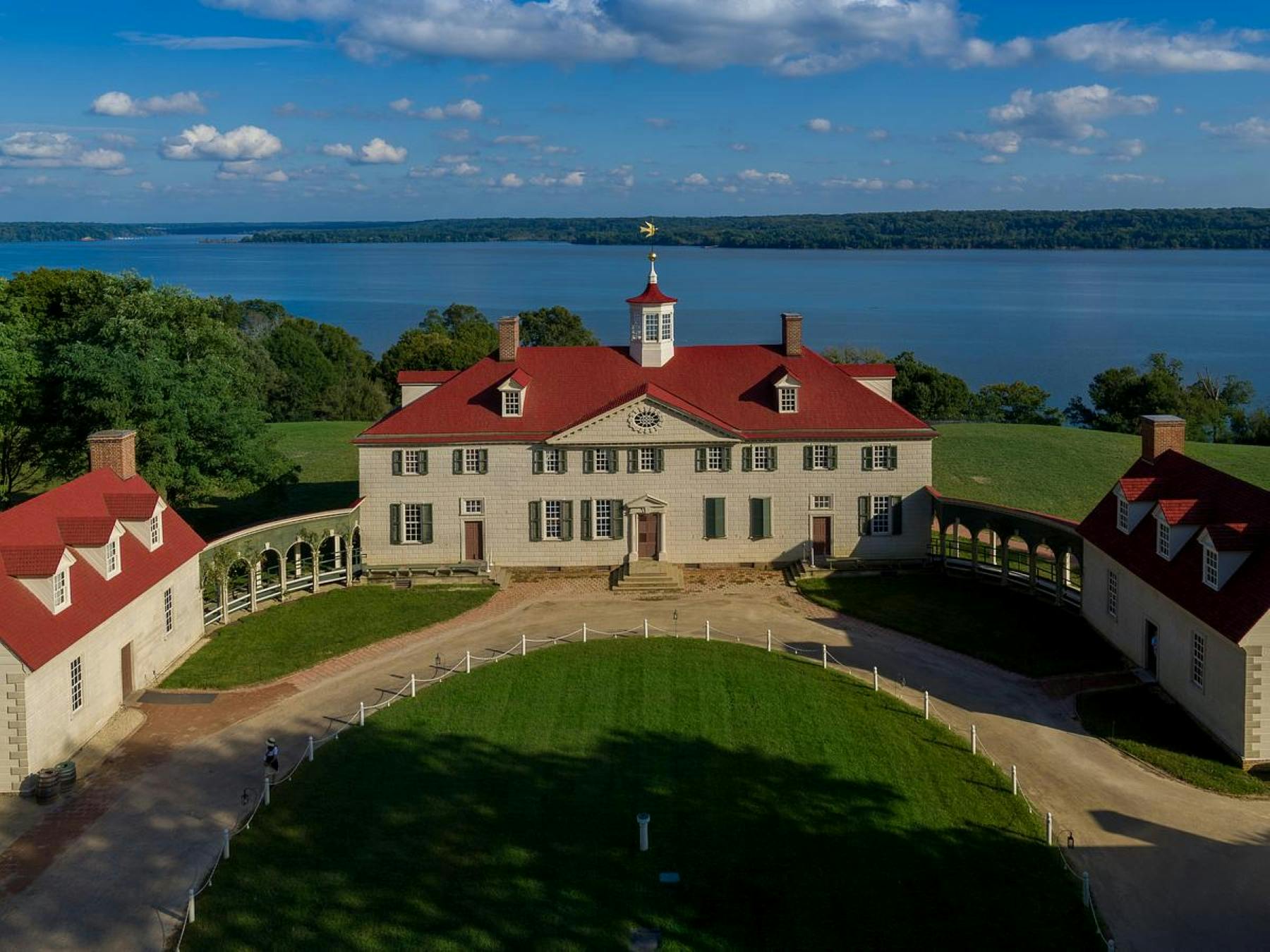 A large house with a red roof and cupola, surrounded by green lawns, overlooks a wide river under a partly cloudy sky.