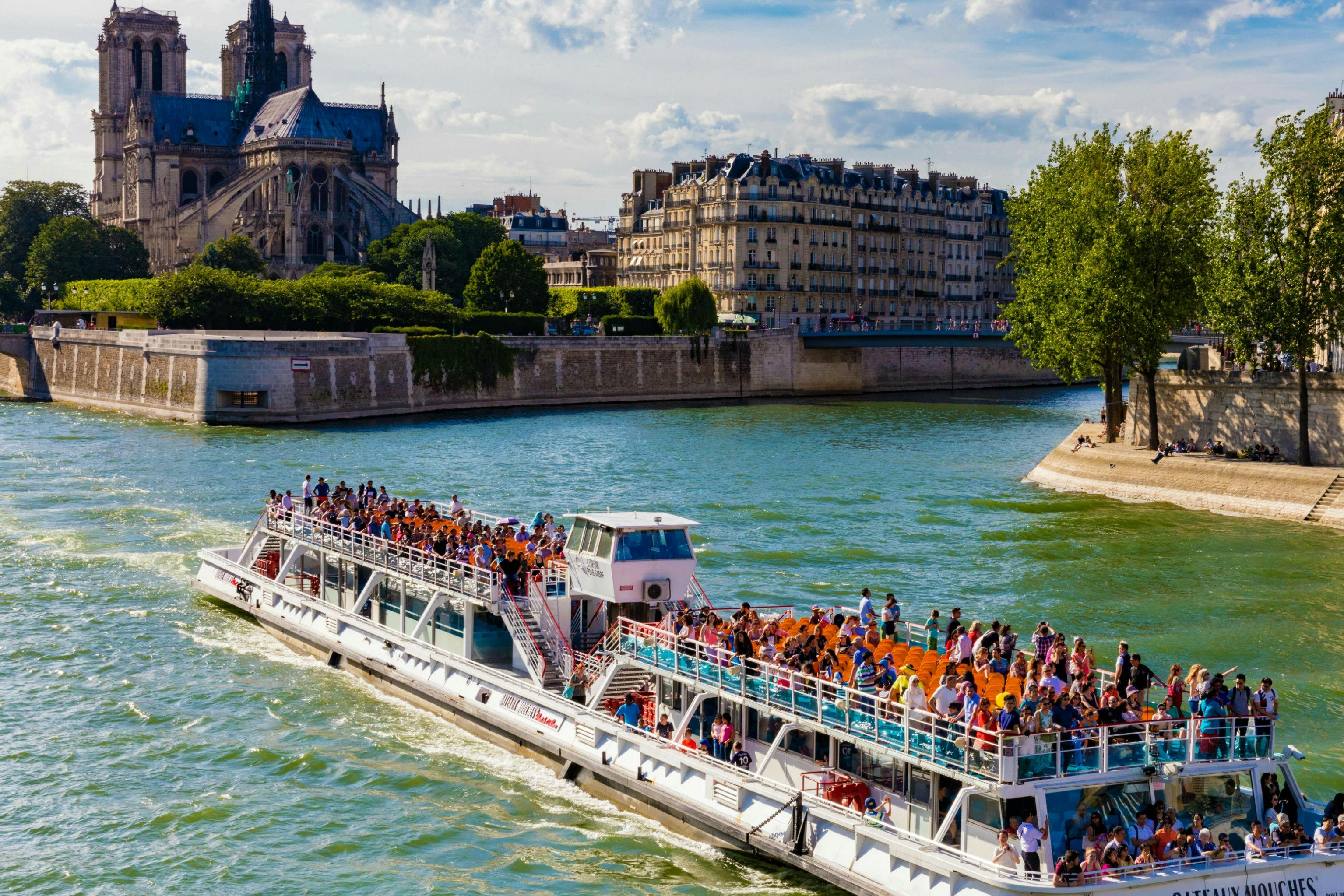 A crowded tour boat travels on a river near historic buildings and trees under a partly cloudy sky.