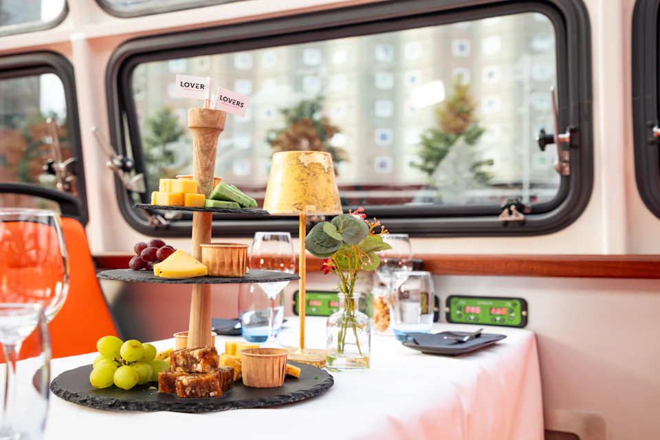 A table set with a tiered tray of cheeses, fruits, and snacks, a vase of flowers, and a golden lamp, inside a vehicle.