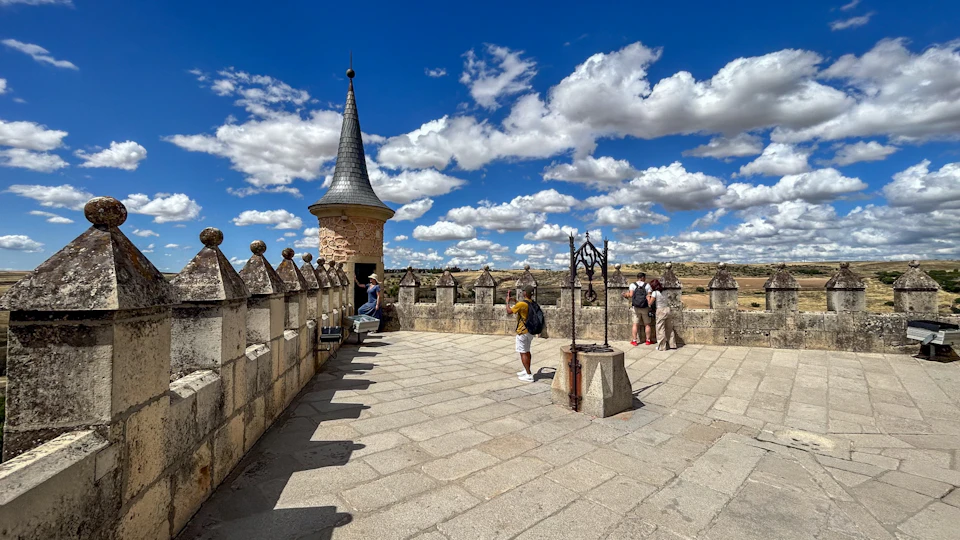 People on a castle terrace with stone battlements, a conical tower, and an iron well structure, under a blue sky with clouds.