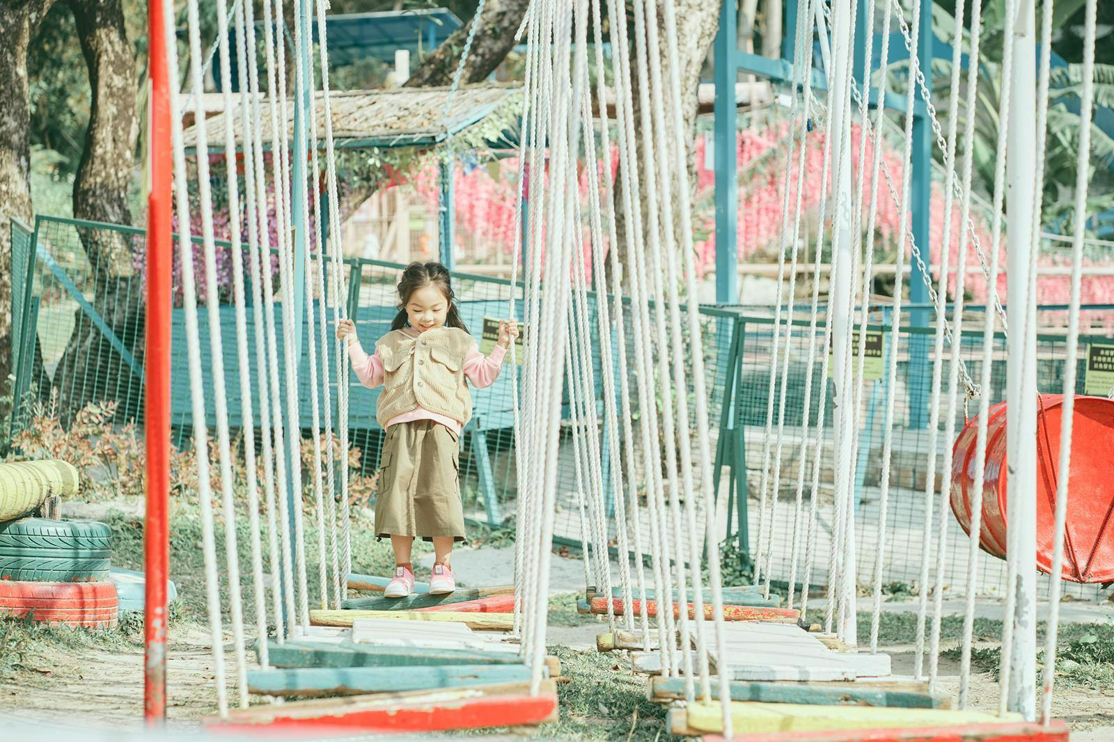 A child crossing a rope bridge in a park, holding onto ropes for balance.