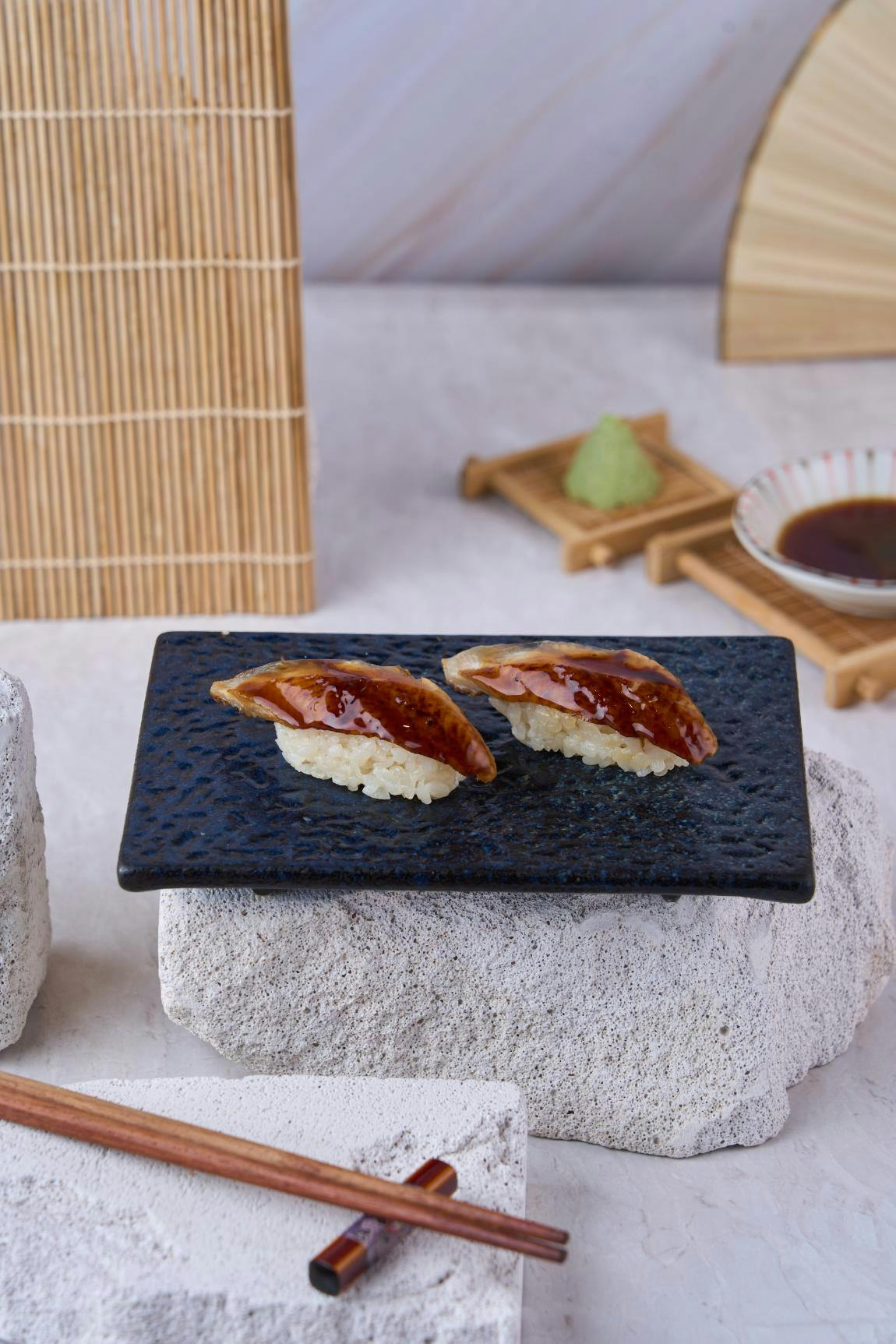 Two pieces of sushi topped with a glazed topping on a black plate, with wasabi and soy sauce in the background.