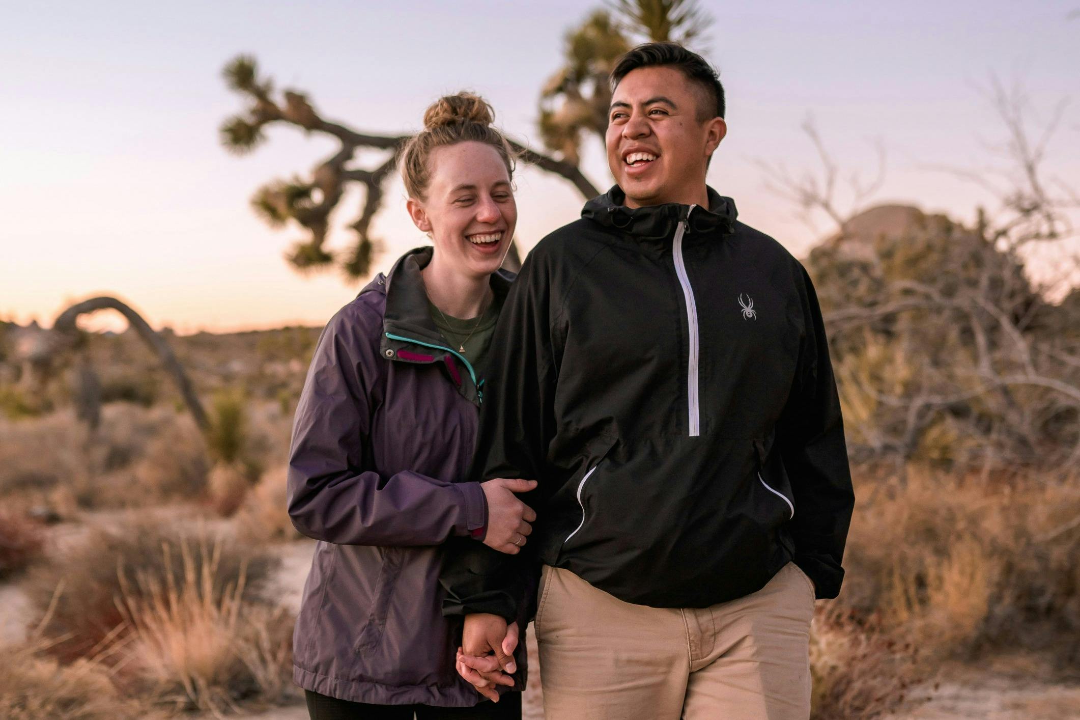 Two people smiling and walking outdoors in a desert landscape during sunset, with Joshua trees in the background.