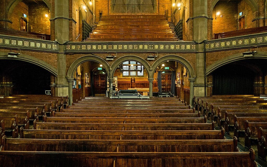 Ornate church interior with wooden pews, arched columns, upper balcony, and lit from wall-mounted lamps.