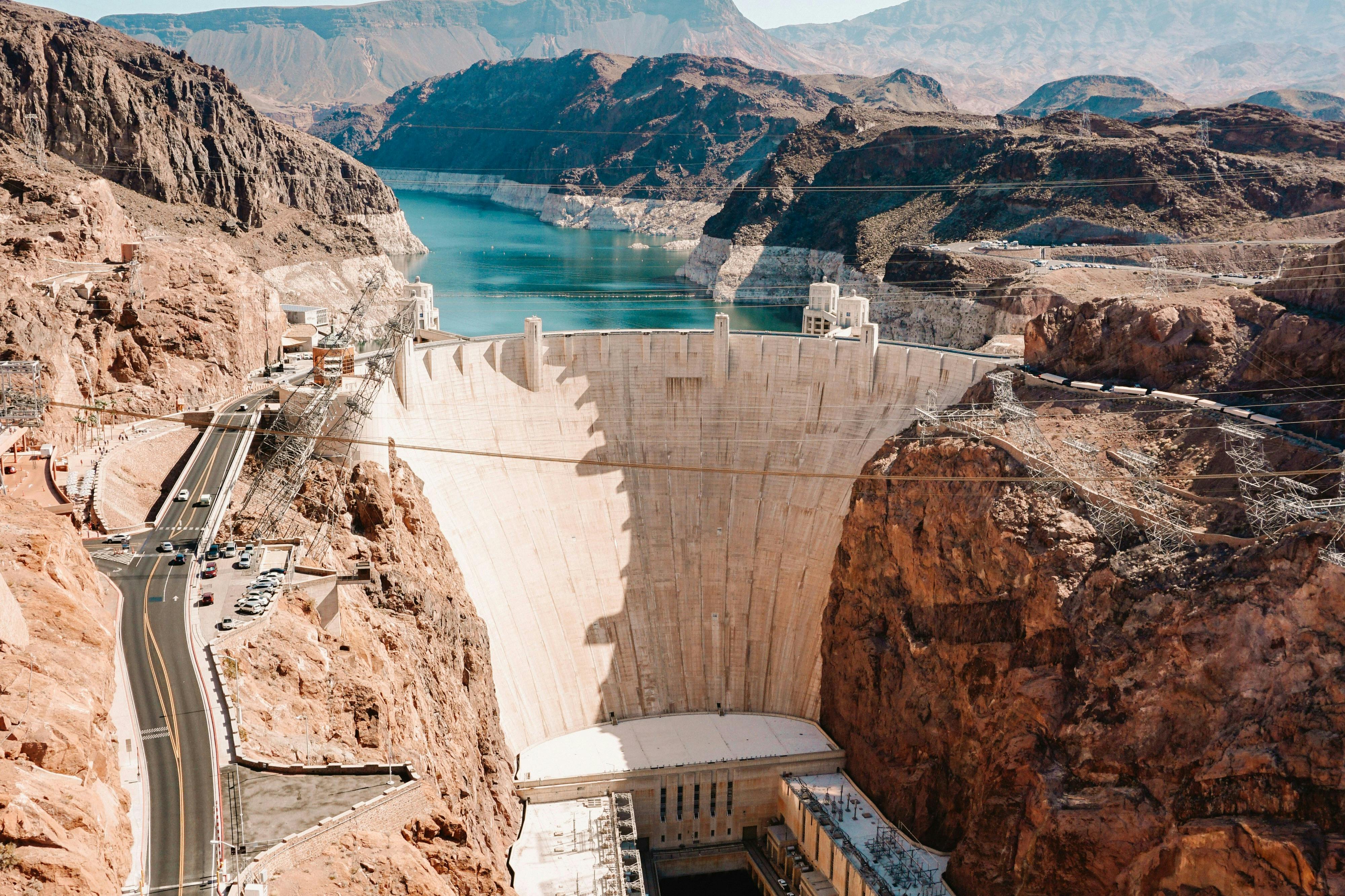 A view of a large dam surrounded by rocky terrain, holding back a reservoir with mountains in the background.