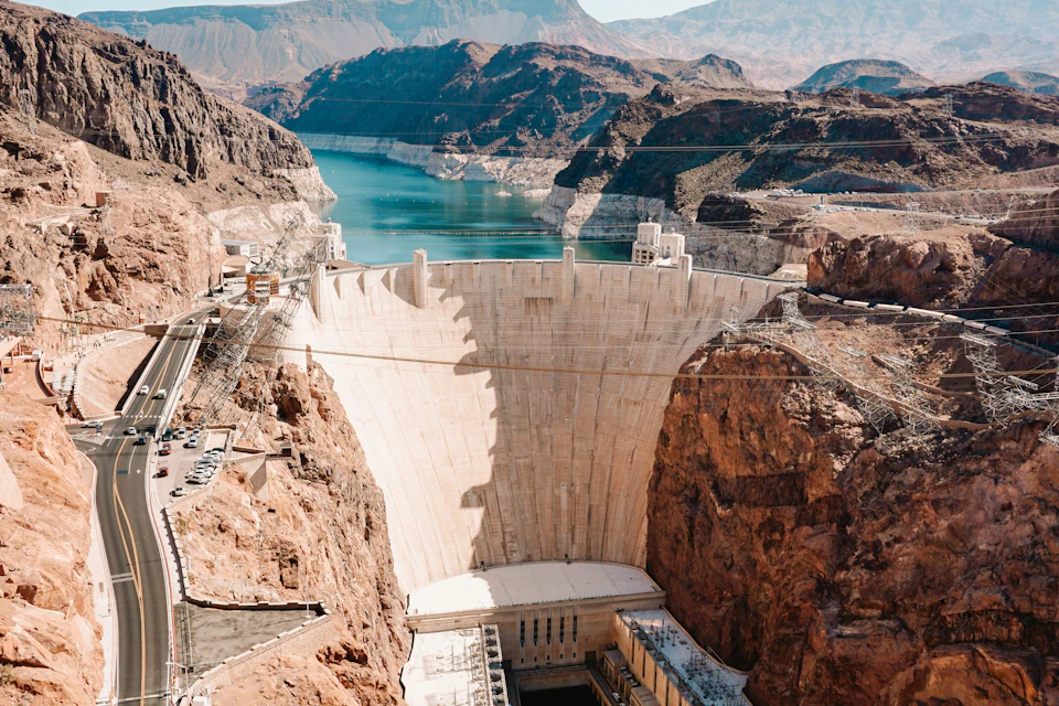 A view of a large dam surrounded by rocky terrain, holding back a reservoir with mountains in the background.