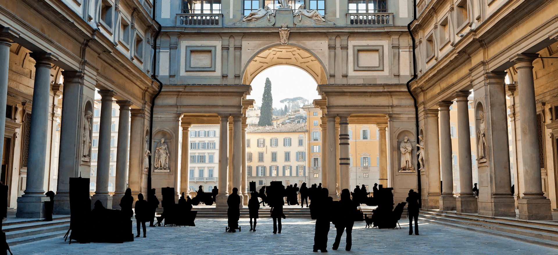 People walk and gather under a large, ornate archway in a historic building with statues and columns. Cityscape visible beyond.