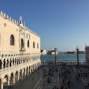 A historical building with arched windows, a busy square, and a waterfront view with distant buildings and boats on the water.
