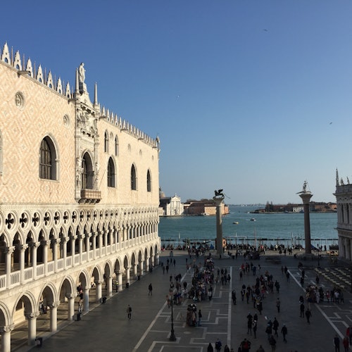 A historical building with arched windows, a busy square, and a waterfront view with distant buildings and boats on the water.
