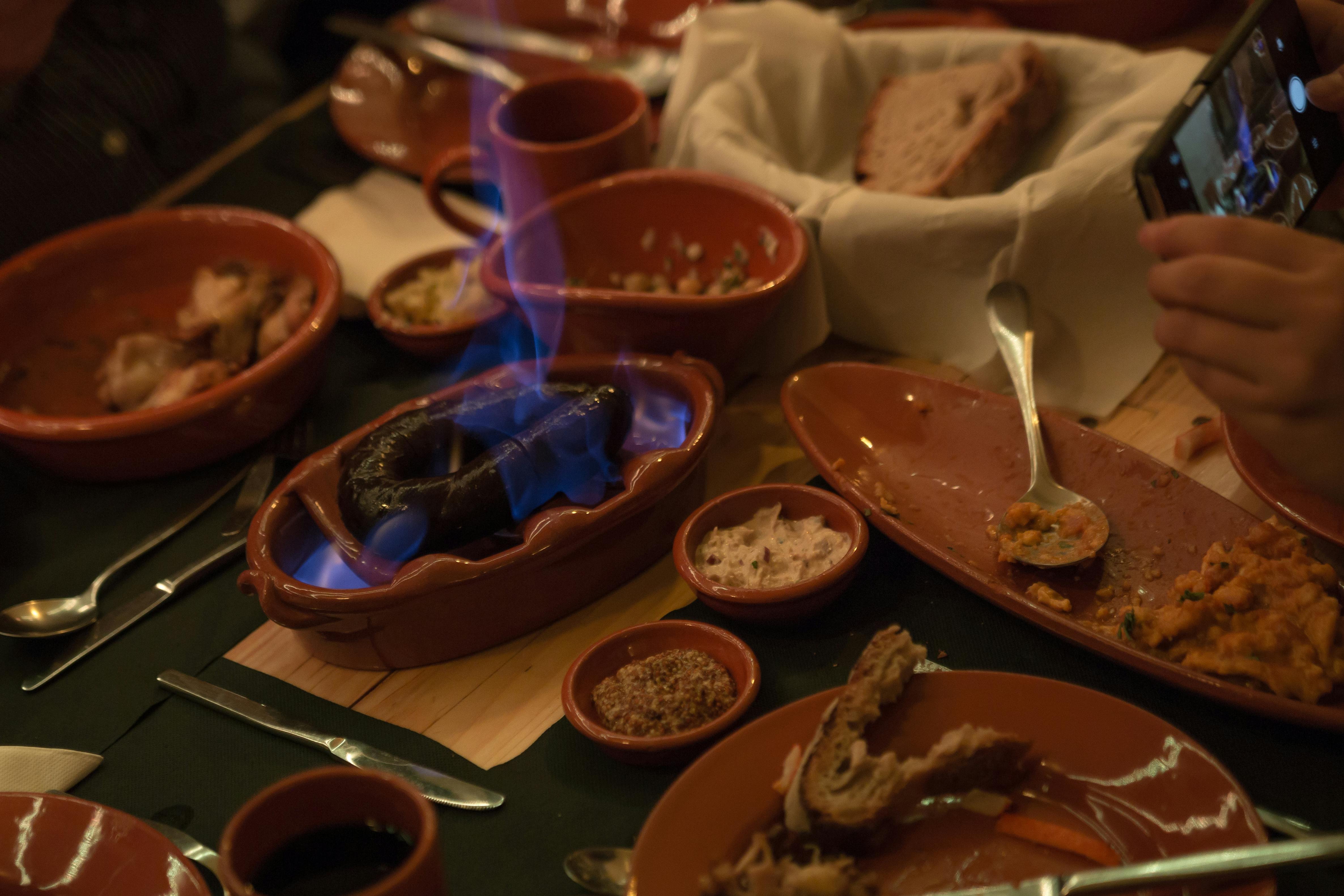 Various clay dishes filled with food, featuring a burning sausage with blue flames on a crowded wooden table.