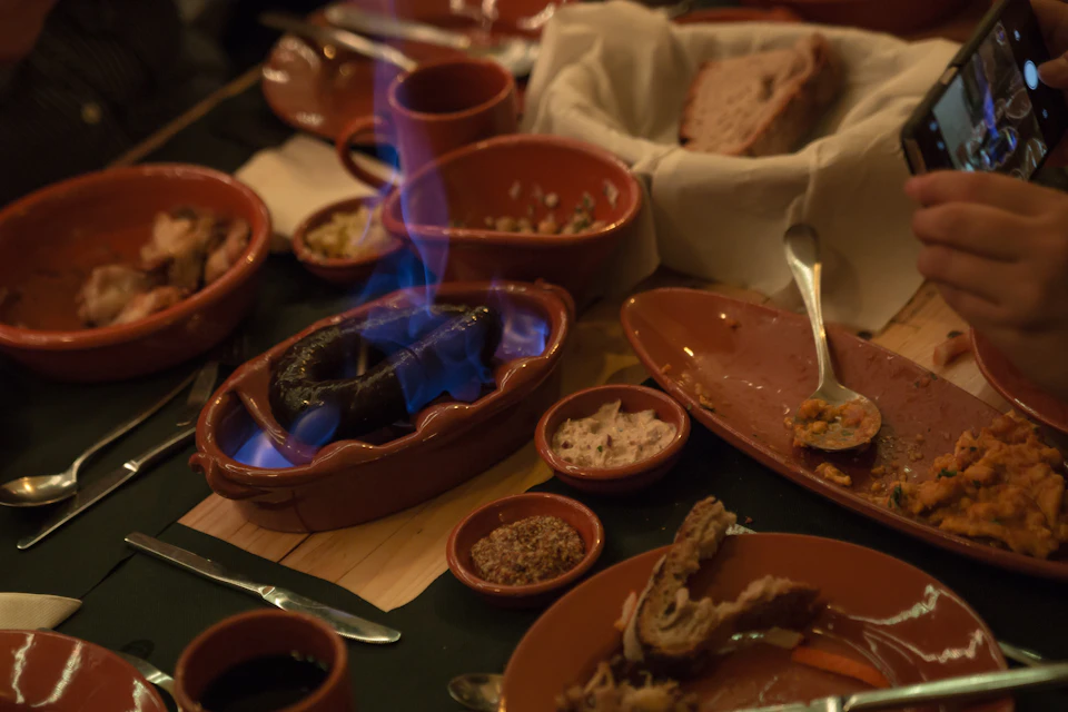 Various clay dishes filled with food, featuring a burning sausage with blue flames on a crowded wooden table.