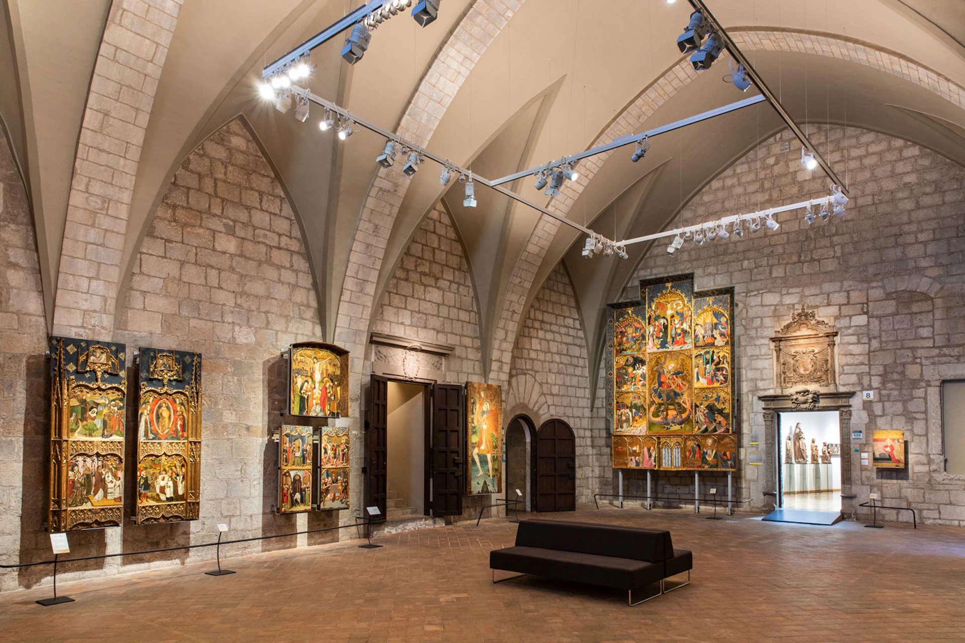 An art gallery with arched ceilings displaying large medieval religious paintings on stone walls with a black bench in the center.