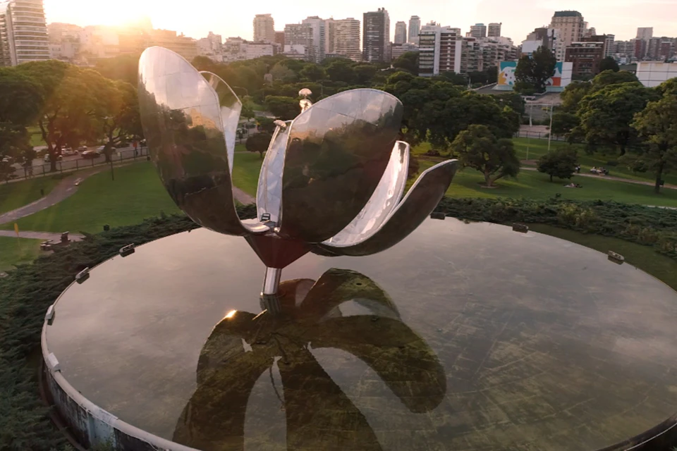 A large metallic sculpture of a flower stands in a reflecting pool, surrounded by a park and urban buildings in the background.