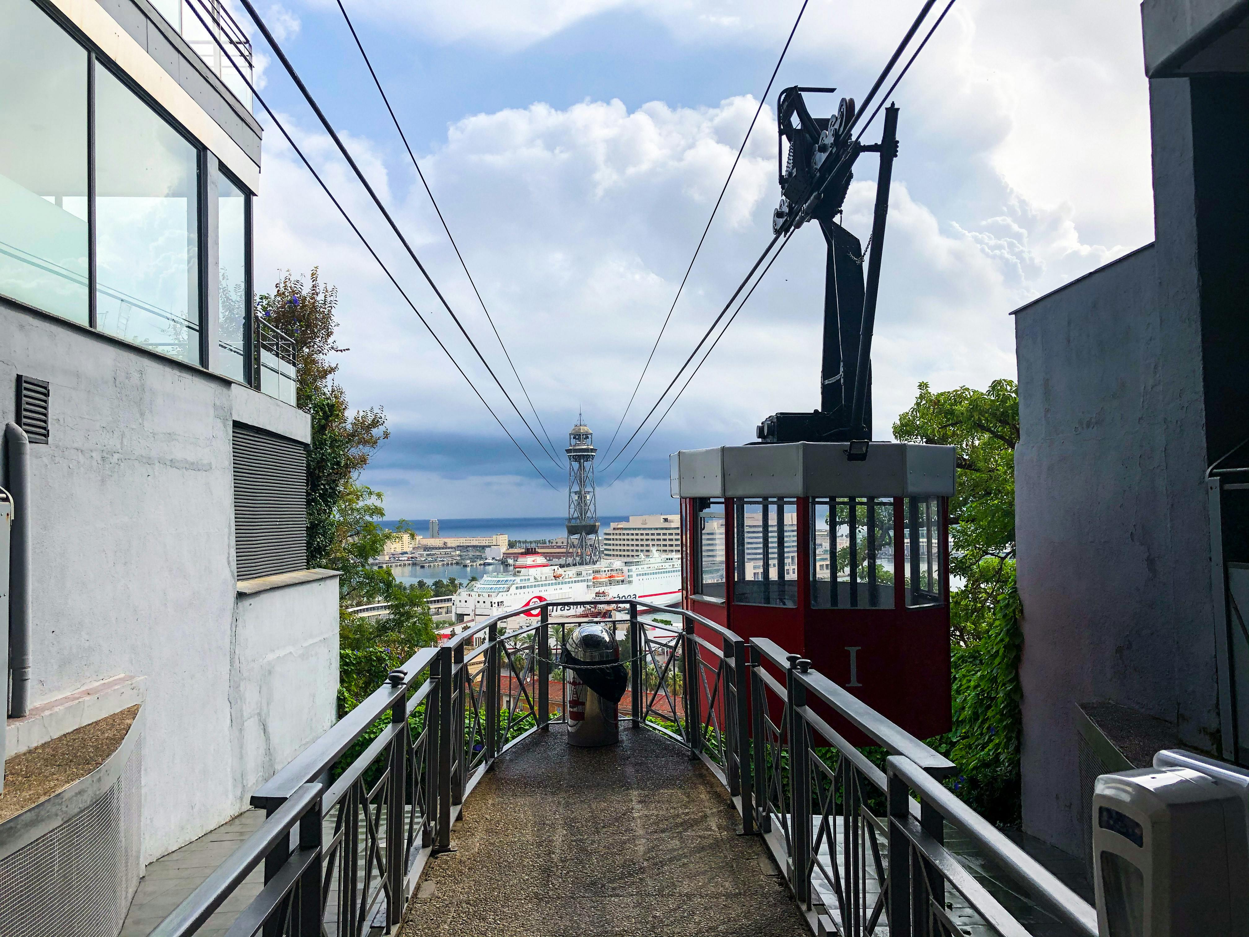 Červená lanovka je zavěšena na drátech nad chodníkem s kovovým zábradlím. V pozadí je vidět panoráma města a zatažená obloha.