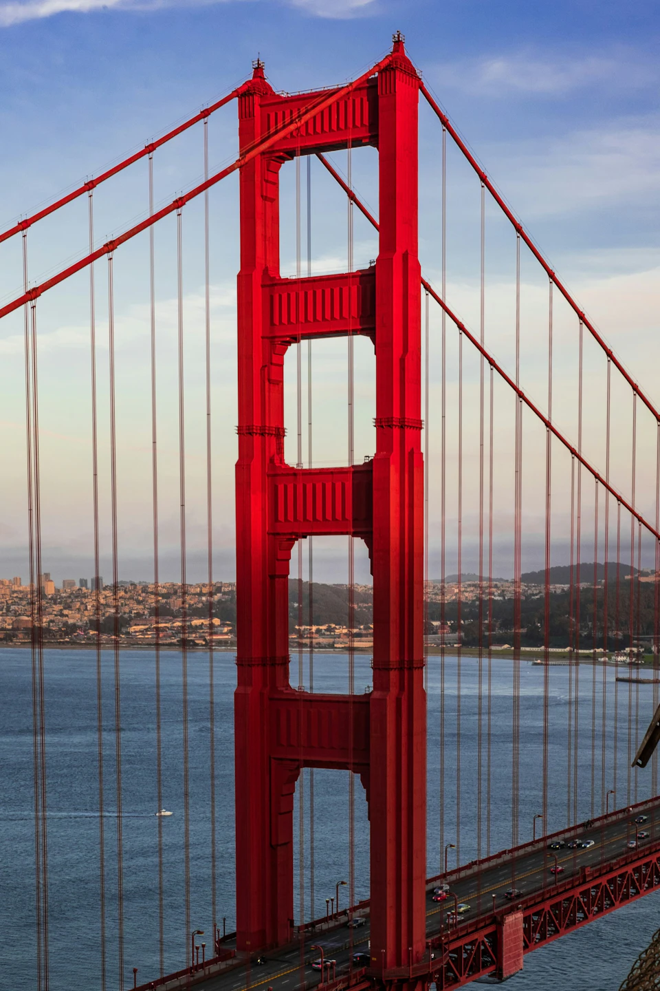 Close-up view of a red suspension bridge tower with cable lines, with a cityscape and water in the background.
