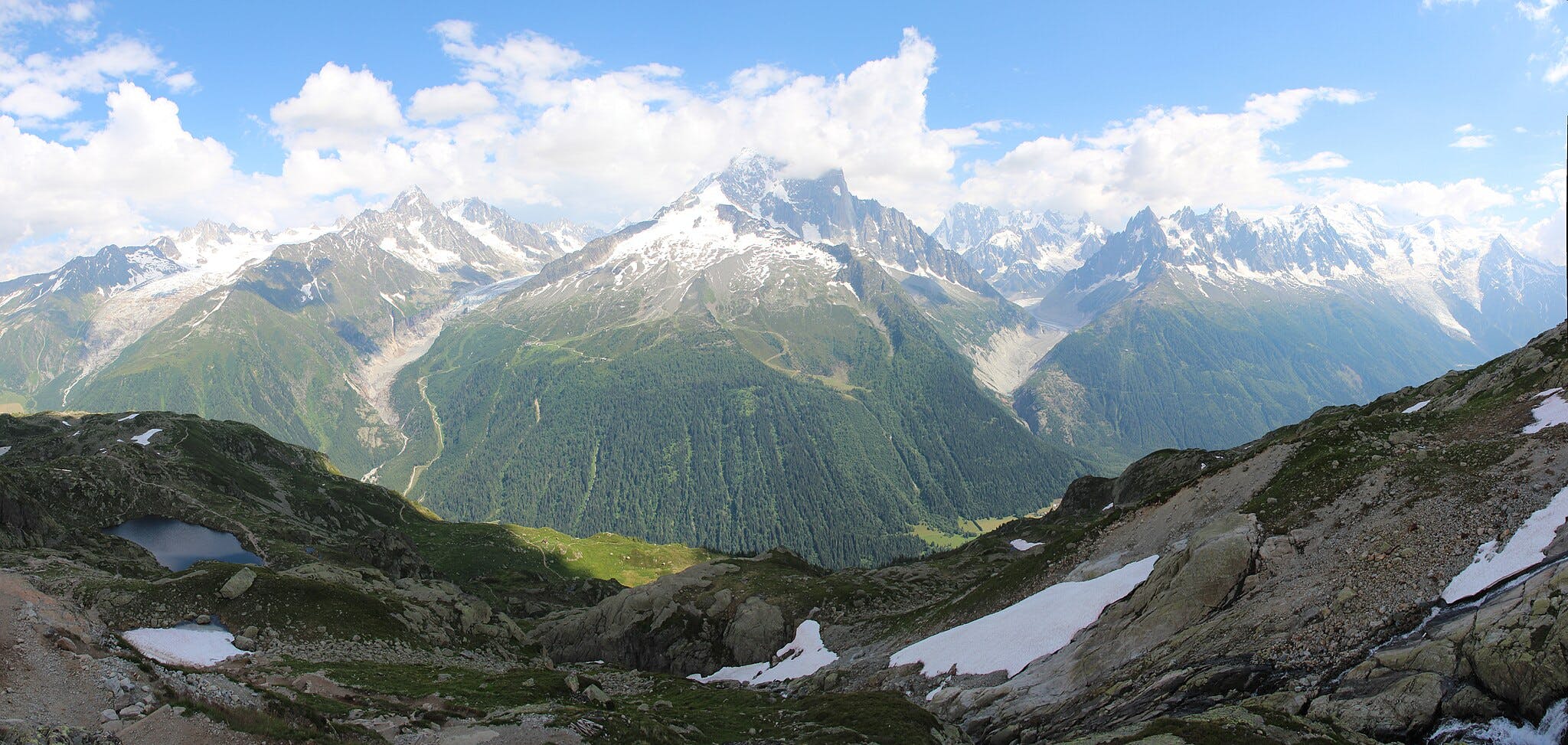 Montagne innevate sotto un cielo azzurro con alcune nuvole, primo piano visibile di un terreno roccioso con chiazze di neve e un piccolo stagno.