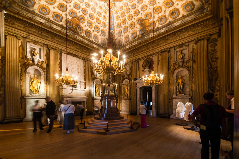 An ornate room with chandeliers, golden decorations, and statues. Visitors are observing the exhibits on display.
