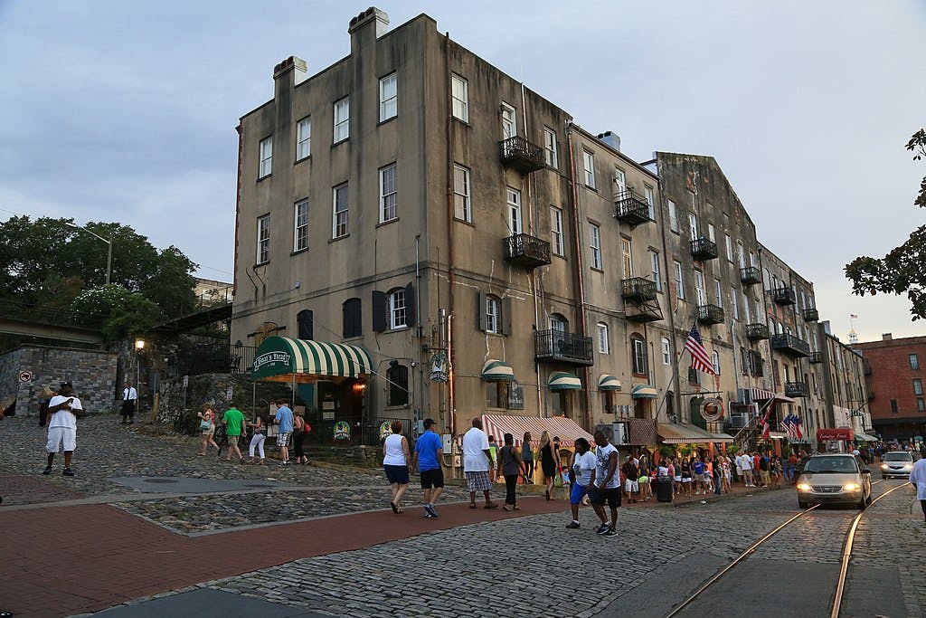 Persone che camminano su una strada acciottolata vicino a vecchi edifici a più piani con balconi, negozi e bandiere americane sotto un cielo nuvoloso.