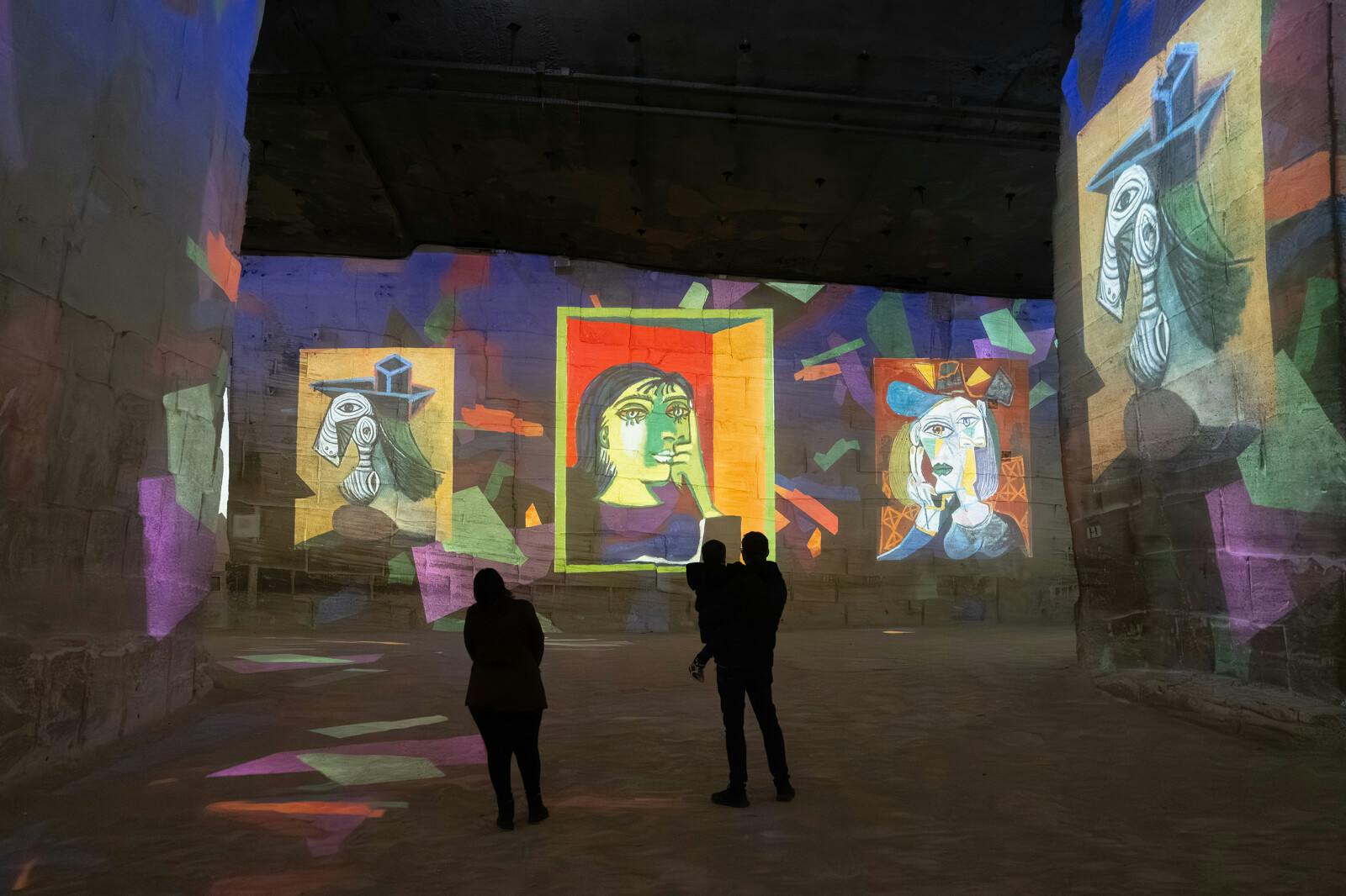 Silhouettes of people standing in a dimly lit room with colorful artworks projected on large walls.