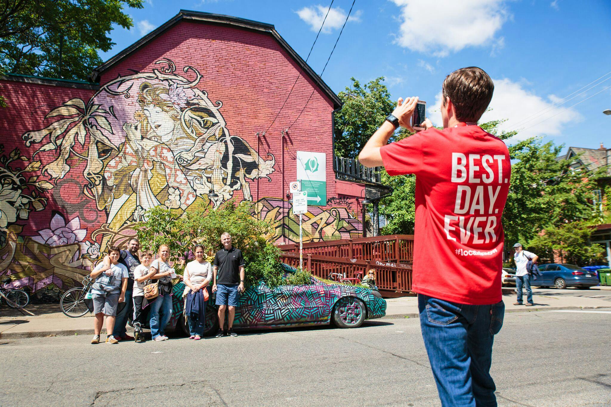 A group of people pose near a colorful car in front of a building with vibrant graffiti art. One person stands off to the side.