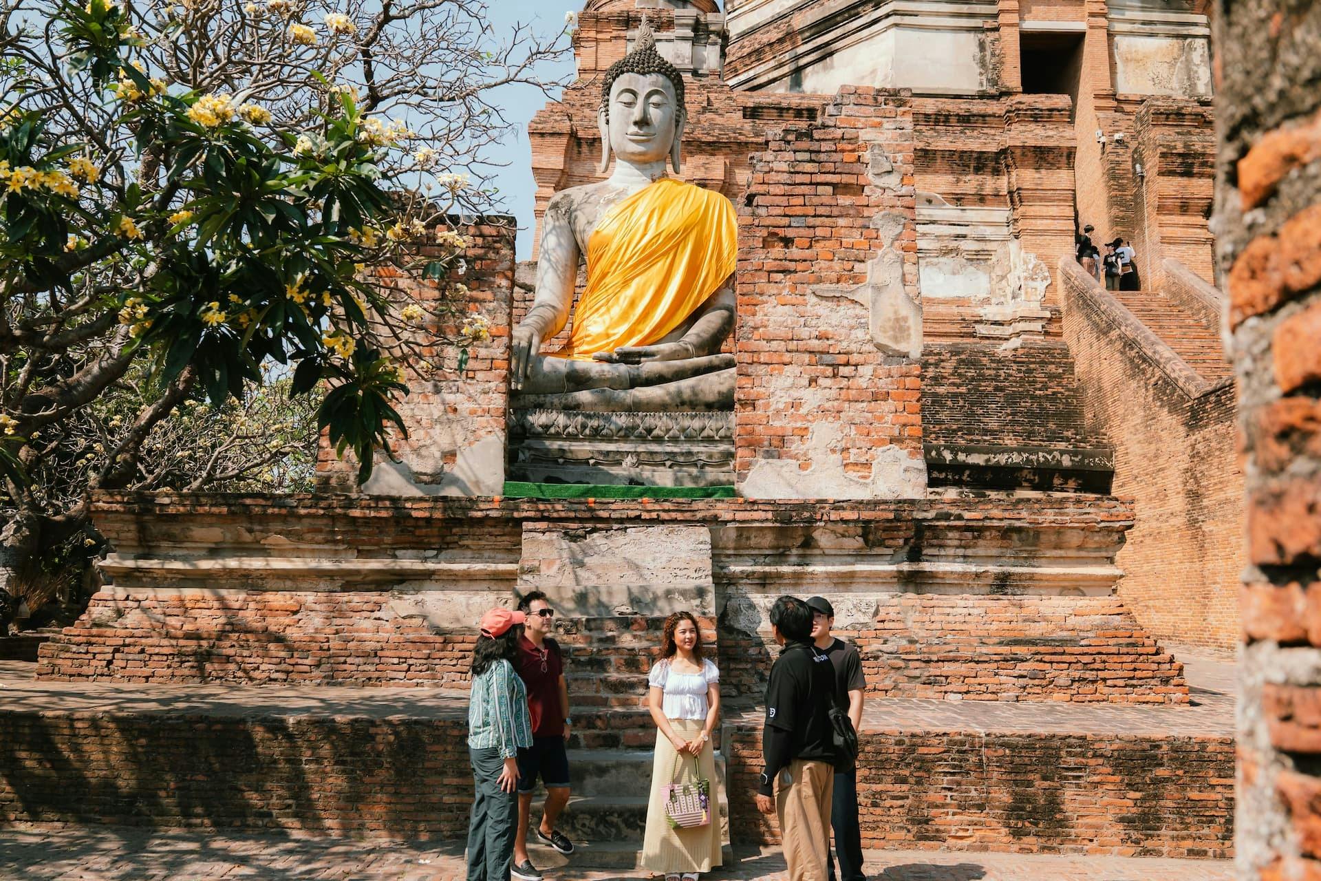 En gruppe mennesker står i nærheden af en stor Buddha-statue draperet i gult, opstillet mod en gammel murstensstruktur og træer.