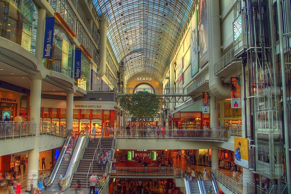 A spacious mall interior with multiple levels, shops, a glass roof, escalators, and numerous people walking and shopping.