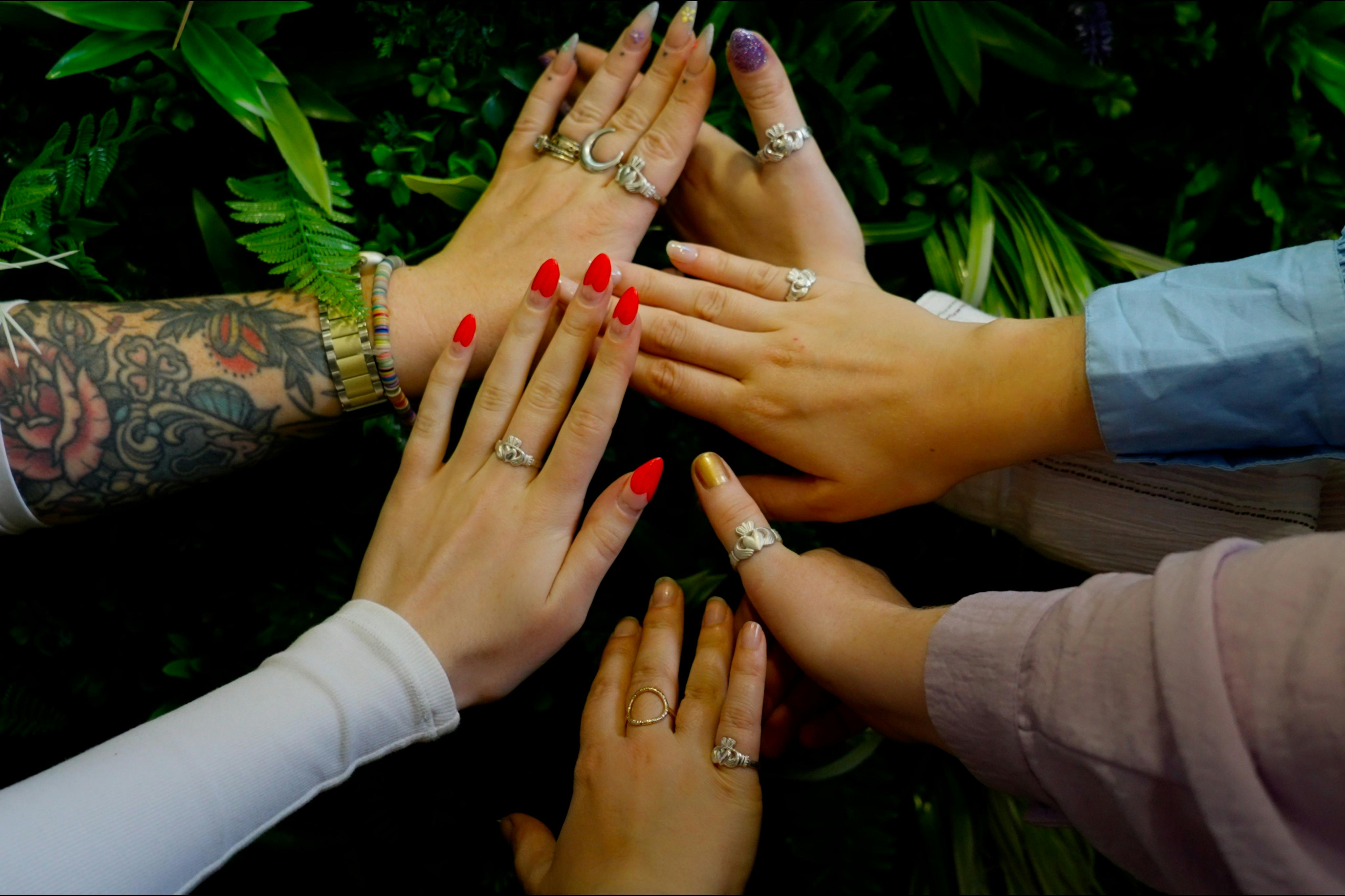 Five hands with varied rings and nail colors, extended in a circle over a backdrop of green leaves.
