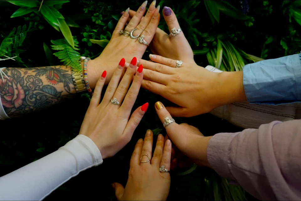 Five hands with varied rings and nail colors, extended in a circle over a backdrop of green leaves.