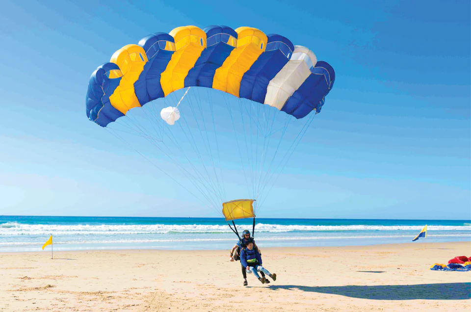 A person with a parachute lands on a sandy beach near the ocean under a clear blue sky.