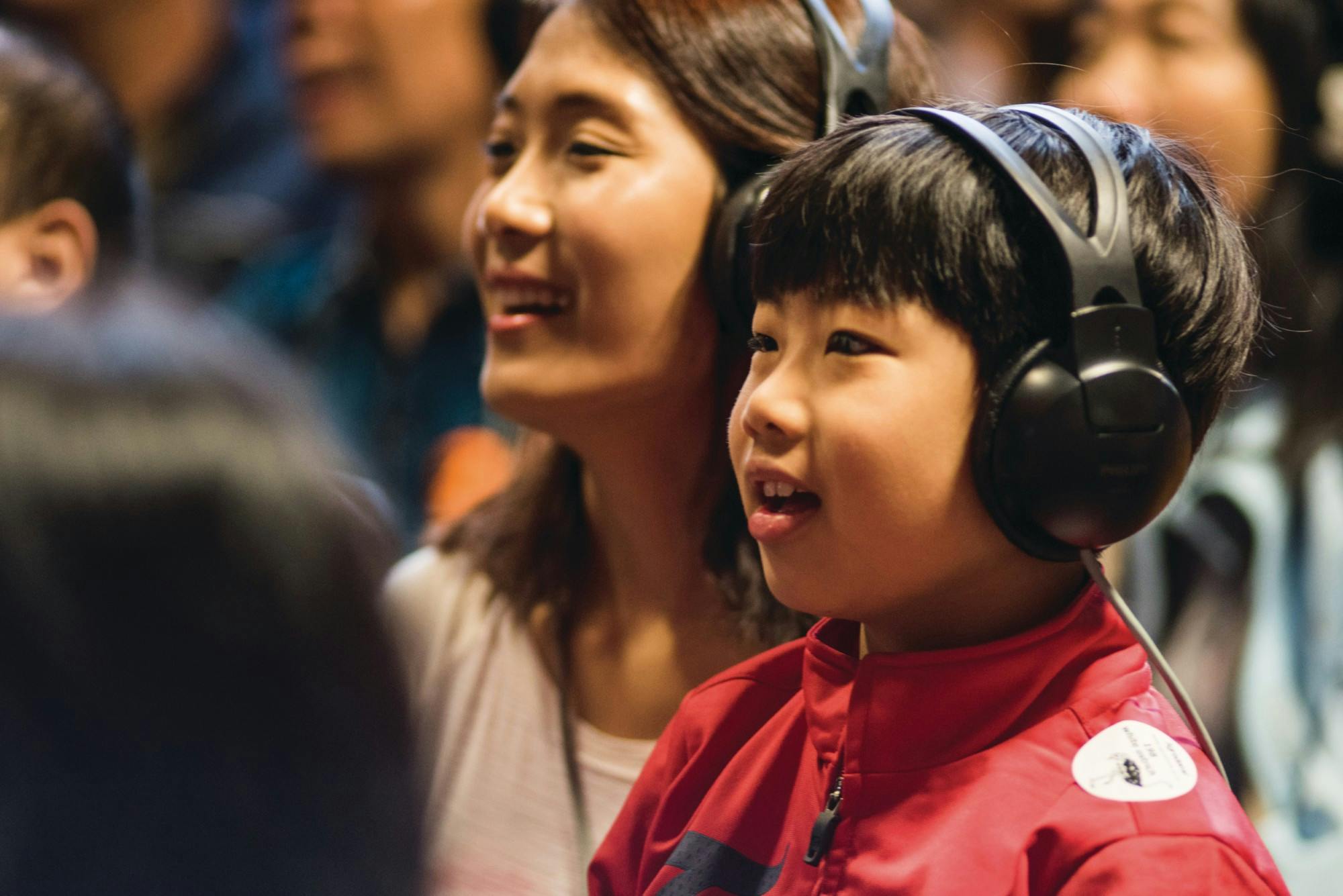 A child wearing headphones and a red jacket sits next to an adult in a lively, indoor setting.