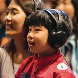 A child wearing headphones and a red jacket sits next to an adult in a lively, indoor setting.