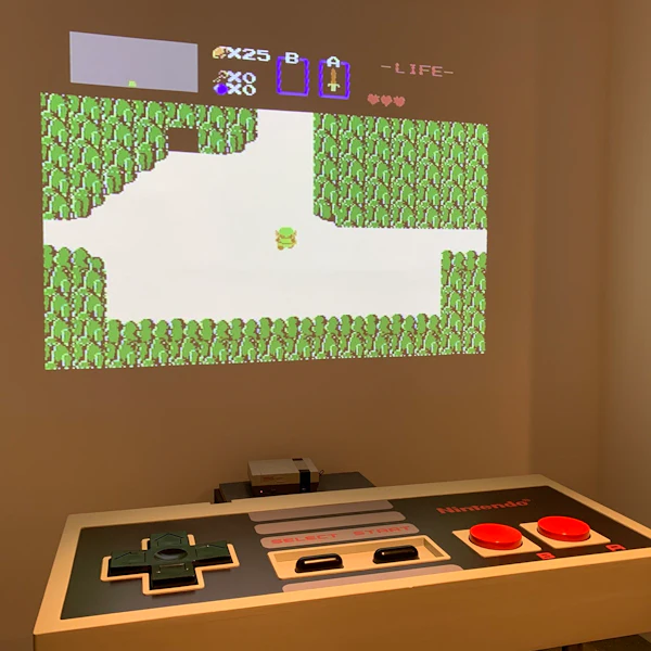 Μια σκηνή από ένα βιντεοπαιχνίδι 8-bit προβάλλεται σε έναν τοίχο, πάνω από ένα μεγάλο χειριστήριο και κασέτα παιχνιδιών της Nintendo.