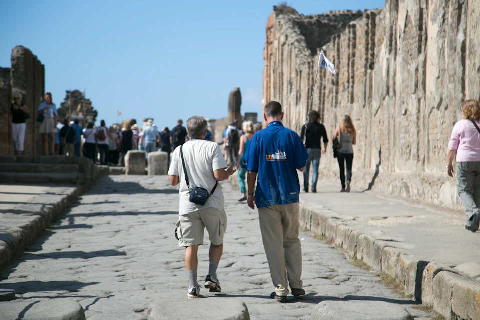 Two men walk on a stone-paved path lined with ancient ruins, heads of a small group visible ahead. A tour guide holds a flag.