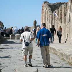 Turister går genom en gammal stengata kantad av förstörda väggar, ledd av en guide som håller en flagga under en klarblå himmel.