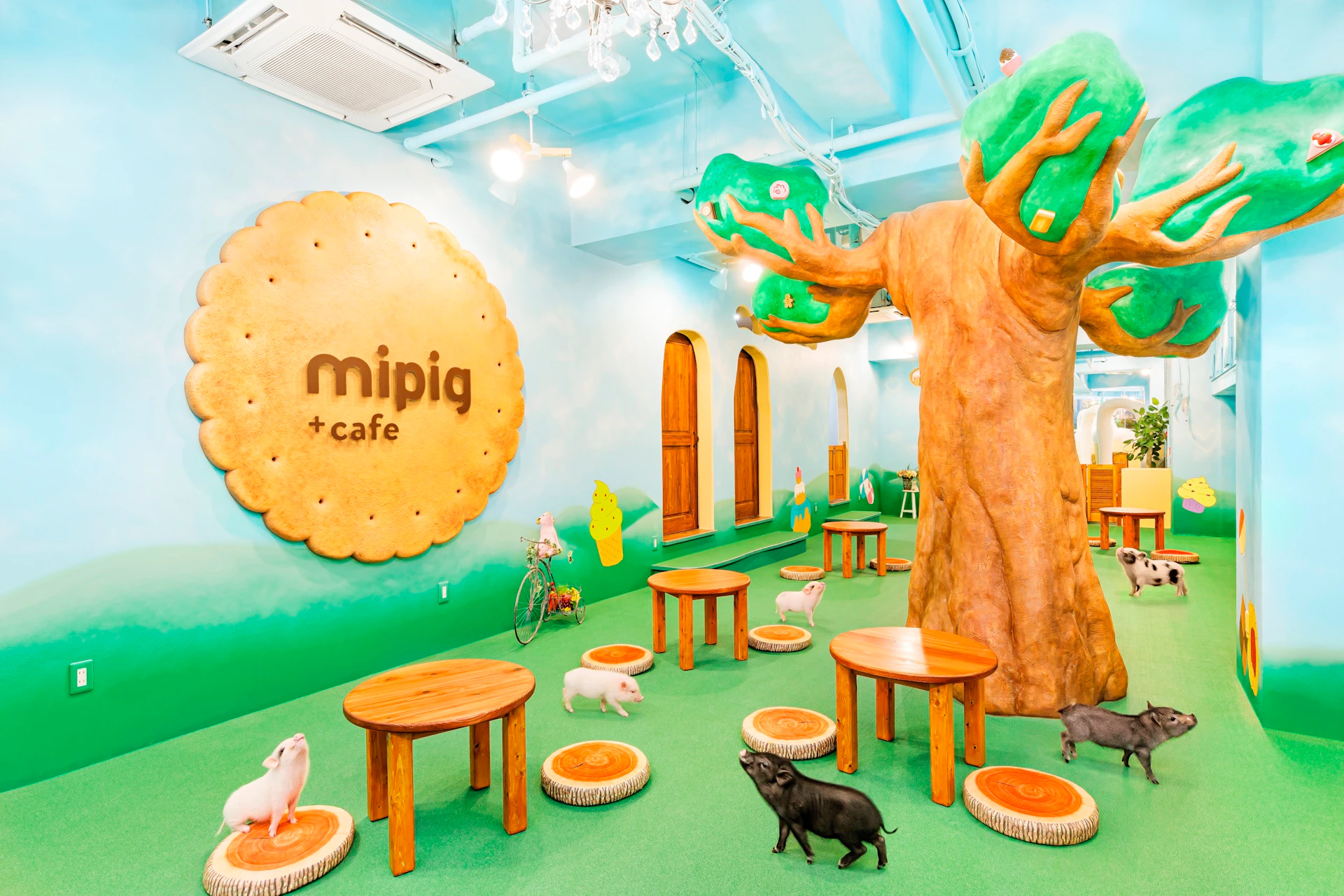 A themed cafe with piglets roaming on a green floor, round wooden tables, a tree-shaped structure, and whimsical decor.