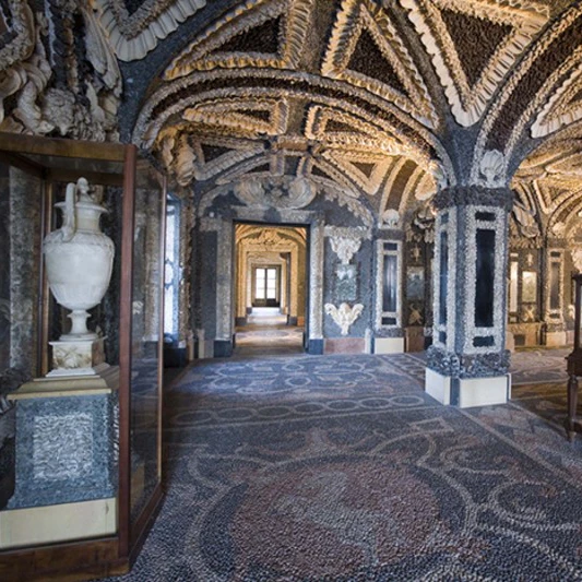 Intricate room with ornate ceiling and walls, mosaic floor, glass display case holding a white vase, and light streaming through windows.