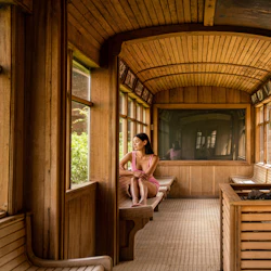 En kvinna sitter på en träbänk i en gammal tågvagn och tittar ut genom fönstret. Bilen har interiör med träpaneler och stora fönster.