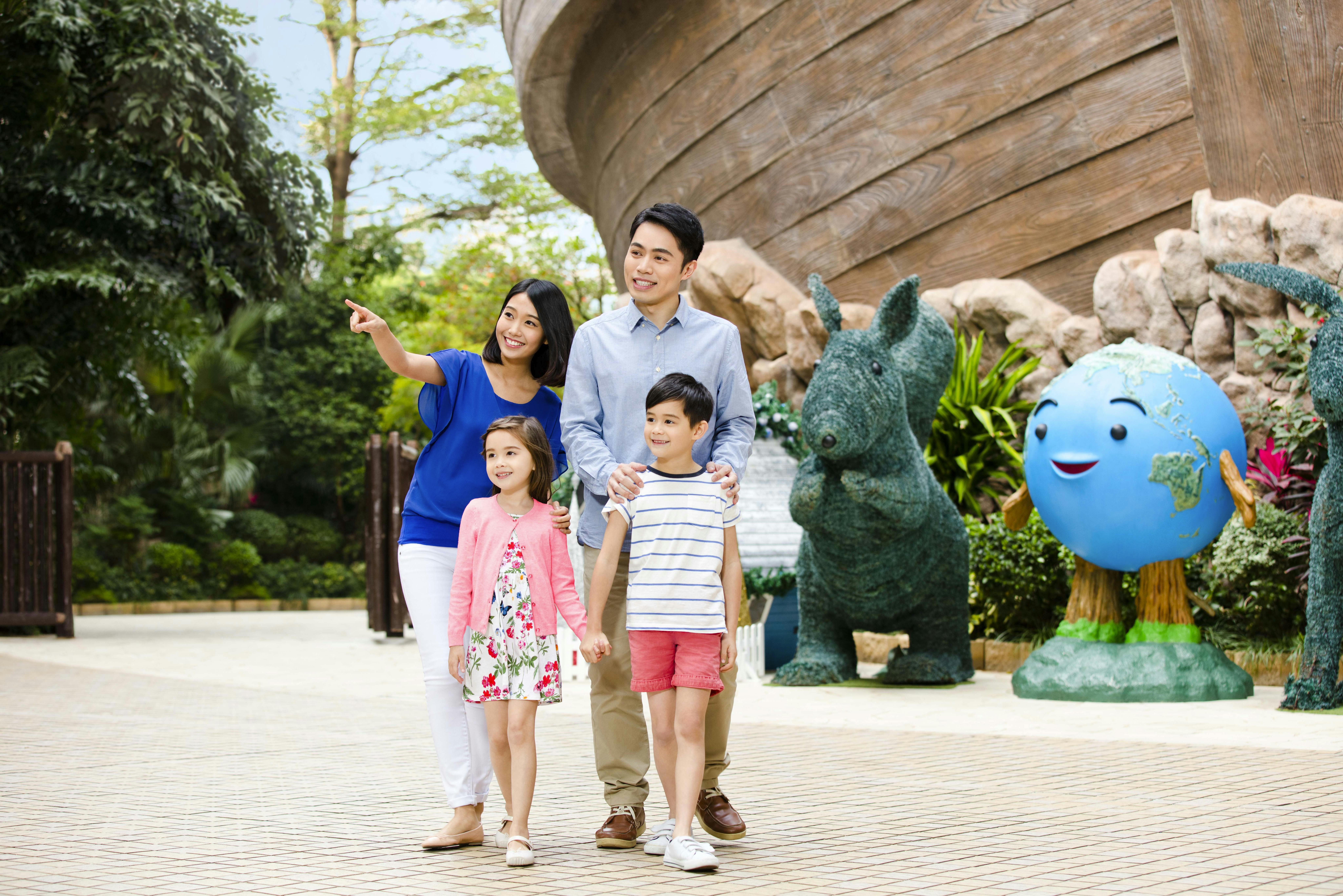 Улыбающаяся семья из четырех человек гуляет на свежем воздухе возле большой статуи зеленого животного и деревянного сооружения, окруженного зеленью.