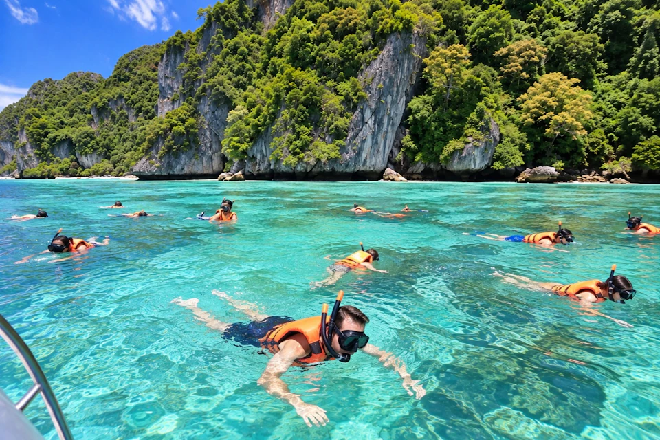 People snorkeling in clear turquoise waters near a lush, rocky island under a sunny sky.