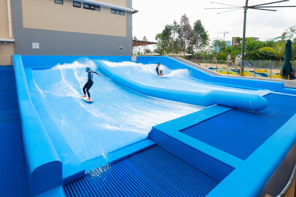 两个穿着潜水服的人在外面的平行人造波浪池上冲浪，后面是一栋现代建筑和树木。