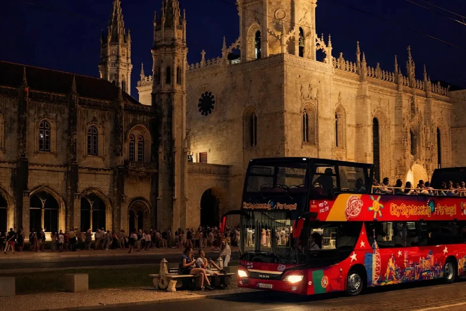 A red double-decker tourist bus in front of an illuminated historic building at night with people nearby.