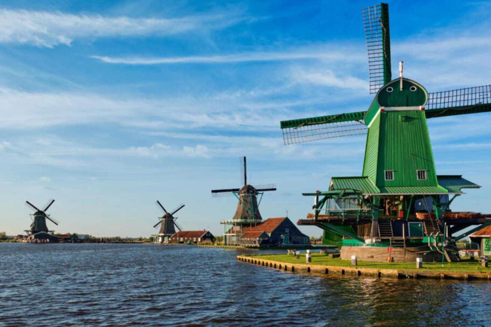 Three windmills near a calm water body under a clear blue sky. Two are in the distance, and one is prominently green and closer.