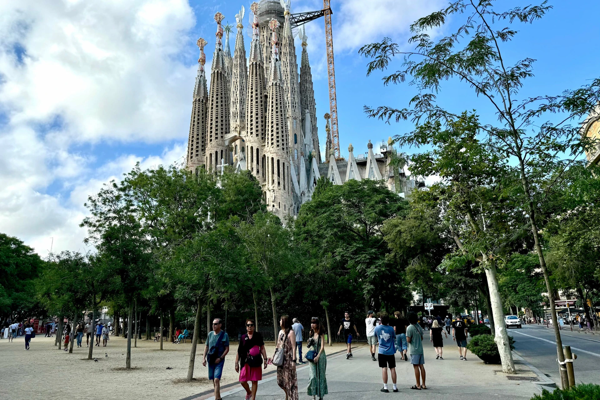 People walking in a park near the Sagrada Família basilica, surrounded by trees, under a partly cloudy sky.