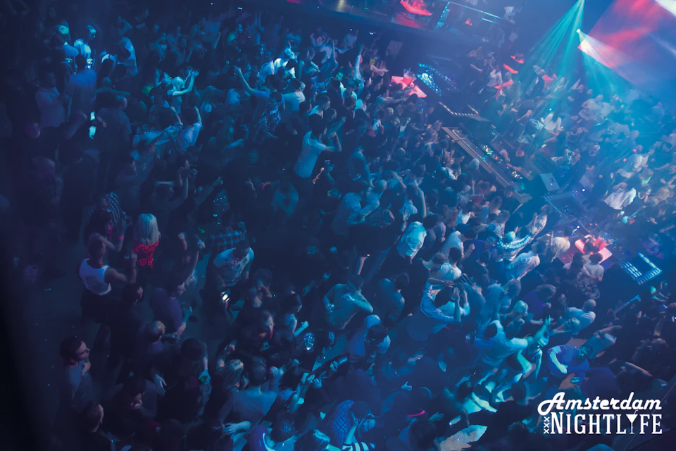Crowded nightclub dance floor with people dancing under blue and purple lights, and a DJ booth visible in the background.